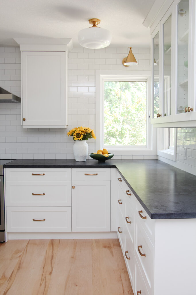timeless white kitchen cabinets, black quartz counter, gold sconce over window, sunflowers and lemon decor, subway tile to ceiling
