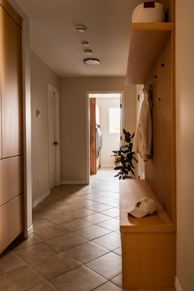 View down mud hall showing custom millwork bench, slat wall detail, and tiled flooring retained for budget conscious renovation.