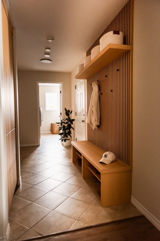 View down mud hall showing custom millwork bench, slat wall detail, and tiled flooring retained for budget conscious renovation.