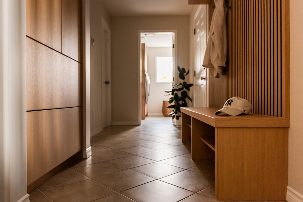 View down mud hall showing custom millwork bench, slat wall detail, and tiled flooring retained for budget conscious renovation.